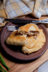Clay plate of fried meat pies with cutlery and green onion on wooden table.
