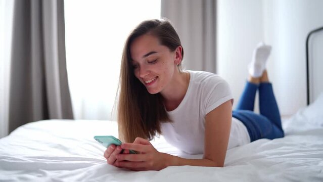 Young happy attractive cute smiling joyful millennial girl lying on a bed and using phone for browsing web, social networking and online chatting at home