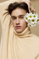 Obraz na płótnie Canvas Close-up photo.A handsome young man with fair skin with dark, short, beautiful hair combed back in a beige turtleneck with a high neck stands on a dark beige background with a bouquet of daisies near