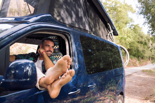 smiling young man relaxing sitting on the seat of his camper van with the feet leaning in the window, concept of nature travel and nomadic lifestyle