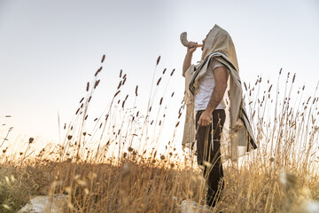 Naklejka premium Jewish man in a traditional tallit prayer shawl blowing the ram's horn shofar, in the field against sunrise sky on Rosh HaShana and Yom Kippurim 