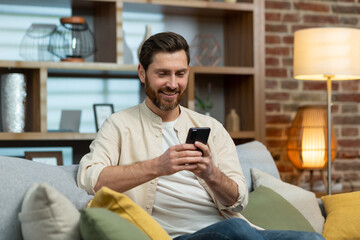 A mature man with a beard in a shirt at home is sitting on the sofa in the living room, using the phone, typing messages and browsing the Internet online.