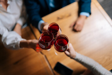 Close-up top view of unrecognizable man and women clinking glasses with red wine during festive dinner at table in luxury restaurant at evening. Happy young friends enjoying nice dinner.