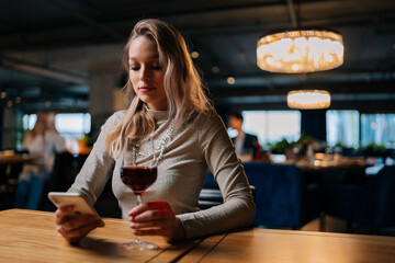 Portrait of serious young woman using smartphone, typing online message sitting at table holding in hand glass of red wine at restaurant with dark interior. Blonde pretty lady chatting with boyfriend.