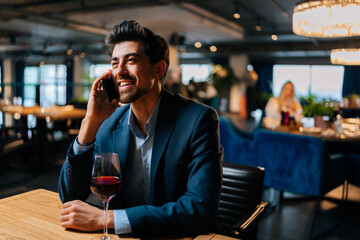 Portrait of happy elegant man wearing fashion suit talking on mobile phone sitting at table with glass of red wine in restaurant. Smiling bearded male waiting for girlfriend on date indoors.