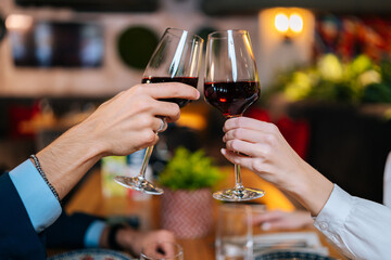 Close-up unrecognizable female and male hands clinking glasses with red wine sitting at table in fancy restaurant at evening. Happy young couple enjoying nice romantic dinner, celebrating anniversary.