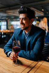 Vertical portrait of cheerful elegant man in fashion suit holding glasses of red wine sitting at table in restaurant, laughing looking away. Bearded businessman resting, having dinner alone.