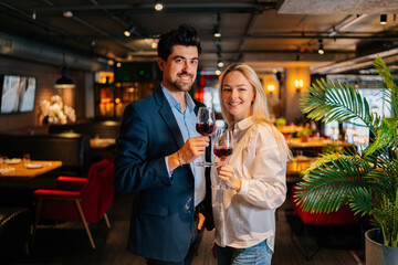 Portrait of smiling couple holding glasses of red wine, looking at camera, standing posing in fancy restaurant at evening. Happy young man and woman enjoying romantic dinner, celebrating anniversary.