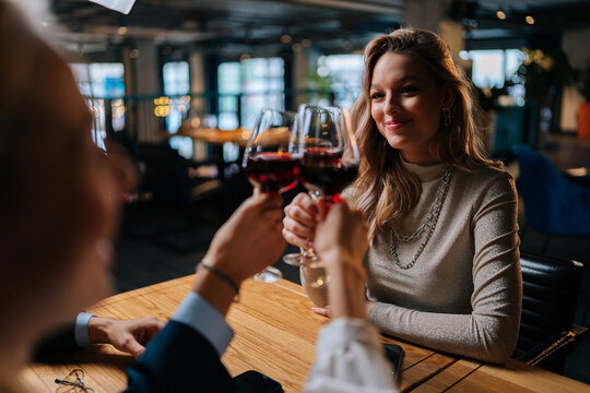 Close-up View From Back Of Unrecognizable Female Clinking Glass Of Red Wine With Cheerful Friends Sitting At Table In Fancy Restaurant At Evening. Happy Three Colleague Enjoying Dinner In Cafe.