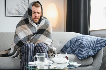 It's cold at home in wintertime. Man freezing in his house in winter because of broken thermostat. young guy wrapped in woolen plaid shivering while sitting on sofa in living room interior