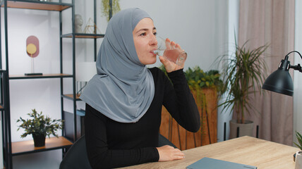 Thirsty young muslim woman in hijab holding glass of natural aqua make sips drinking still water preventing dehydration. Girl at home office with good life habits, healthy slimming weight loss concept