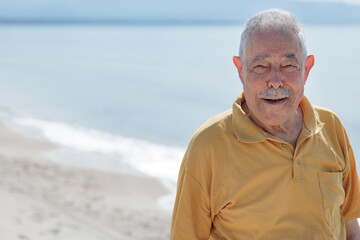 Un signore anziano vestito con polo color senape guarda sorridente  in riva al mare in una spiaggia - obrazy, fototapety, plakaty