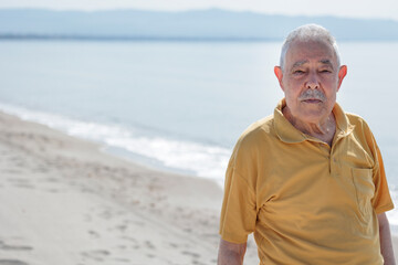 Un signore anziano vestito con polo color senape osserva serio in riva al mare in un apisaggia - obrazy, fototapety, plakaty