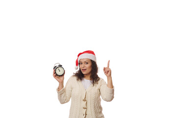 Amazed young brunette pretty woman wearing a Santa hat, holding a black retro alarm clock with...