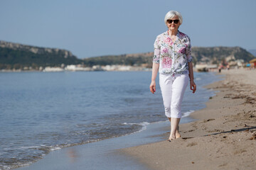 bella signora anziana passeggia in riva al mare, sullo sfondo si vede la spiaggia le montagne - obrazy, fototapety, plakaty