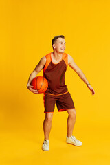 Portrait of young active man in orange uniform training, playing basketball isolated over yellow...
