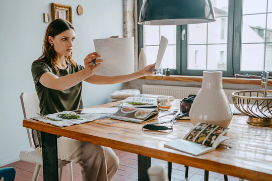 Concentrated female food stylist comparing photographs sitting at table in studio