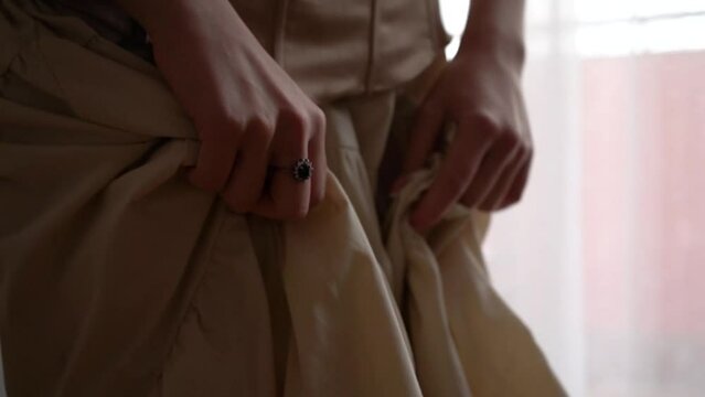 A young woman in a beige retro dress and corset crumples and squeezes the skirt fabric with her hands, an old ring on a girl's hand. Back light, slow motion, close-up details, 4K