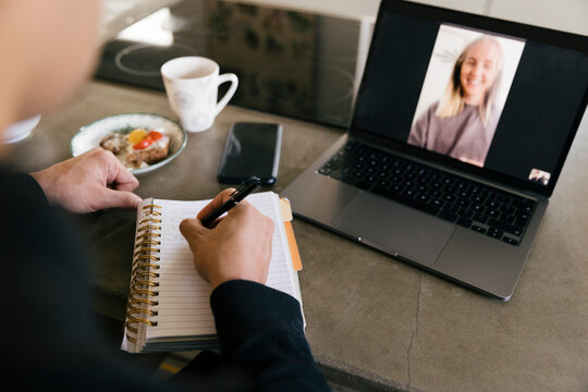Hands of businessman writing in diary while doing video call with female colleague on laptop