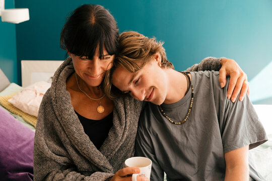 Smiling mother with arm around son leaning on shoulder sitting on bed at home