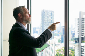 Fototapeta premium Smart Businessman hold tablet and point finger to outsite the window for forecast business trend and cityscape view