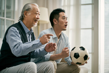 asian senior father and adult son watching football match on tv together