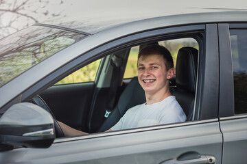 Joyful young man driving safe his new car, looking cheerful to camera. Contented teenage driver enjoying the ride, keeps hands on the steering wheel traveling, road trip concept - 551836335