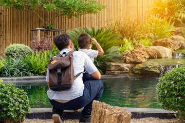 Back view of Asian father wear face mask with backpack embraces his son wear mask to watch carp...