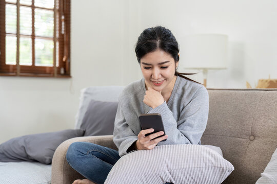 Image of smiling nice asian woman using cellphone while sitting on sofa