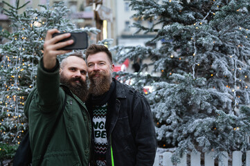 Long beard hipsters homosexuals enjoying Christmas advent, smiling and taking selfie in front of Xmas tree outdoor. Attractive male gay couple celebrating holiday season.
