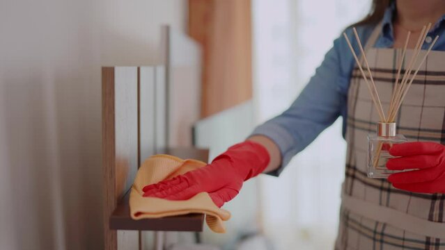 Housewife wearing apron and protective rubber gloves wipes dust with rag from shelf during house cleaning