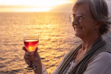 Smiling senior woman with a glass of wine in hand standing in front of the sea while the sun sets in the sea. Peace, solitude, relaxation, happy retirement