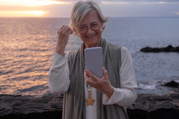 Smiling elderly woman standing in front of the sea looks at her mobile phone while the sun sets in...