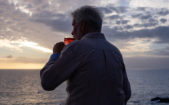 Rear view of senior man sitting in front of the sea at sunset drinking a glass of wine, horizon over the water. Lonely old man drowns his problems in alcohol