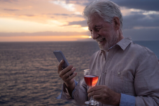 Smiling elderly man with a glass of wine in hand standing in front of the sea looking at mobile phone while the sun sets in the sea. Peace, solitude, relaxation
