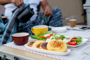 Several types of food for camping traveler include pancake, vegetable salad and coffee are put on table with man pour tea into cup in breakfast.