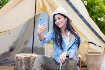 Asian woman is using mobile phone to take selfie photo at her tent while camping outdoor during...