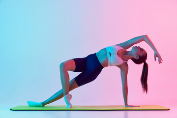 Portrait of young sportive woman training, standing on side plank isolated over gradient blue pink background in neon light. Concept of sport, fitness, health