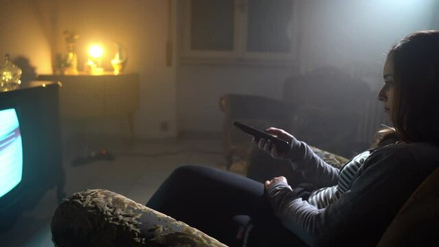 Side view shot of woman sitting in armchair in dark illuminated room and changing TV channels with remote control, sad female searching working broadcast channels on retro television, old broken TV
