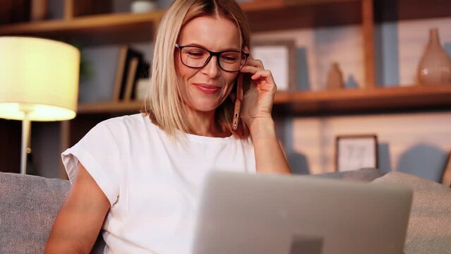 Portrait of busy smiling middle aged woman with eyeglasses talking by phone call conversation while has laptop work in the living room at home Remote work freelance concept