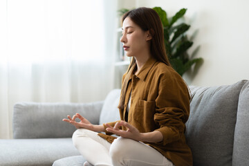 Peaceful asian young woman, girl hands in calm pose sitting practice meditating in lotus position on sofa at home, meditation, exercise for wellbeing, healthy care. Relaxation, happy leisure people.