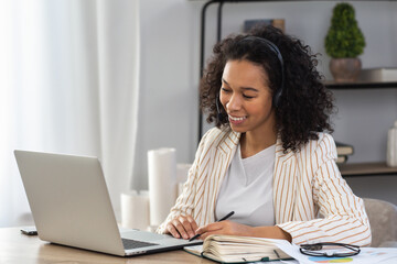 Successful African American woman entrepreneur or office worker in headset using a laptop for video conference with employees sitting at the desk in the office