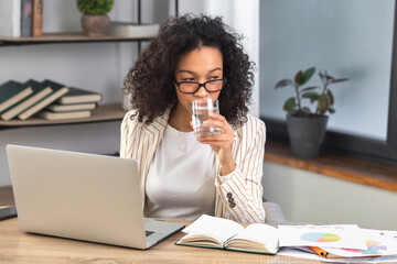 Tired african american business woman taking a sip of clean water sitting at the table in the office. Recuperation concept