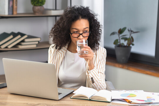Tired african american business woman taking a sip of clean water sitting at the table in the office. Recuperation concept