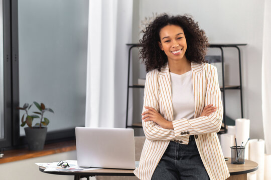Young successful African American woman entrepreneur or an office worker stands with crossed arms near a desk in a modern office, looking at the camera and smiling