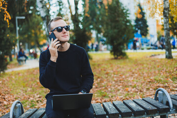 Cute successful young man in sunglasses sitting on park bench with laptop and phone and looking away on sunny day. Freelance, remote work concept.