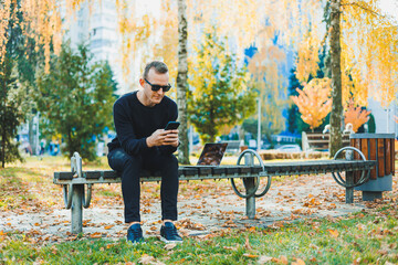 Cute successful young man in sunglasses sitting on park bench with laptop and phone and looking away on sunny day. Freelance, remote work concept.