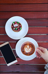 Hand Holding a Cup of Hot Cappuccino Coffee Beside with a Plate of Christmas Chocolate Cupcake and Smartphone