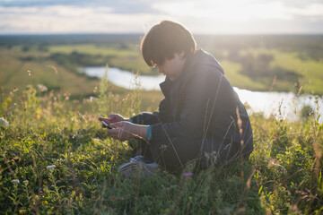 A teenage boy is sitting on the grass on a mountain against the backdrop of a beautiful sunset sky and using a mobile phone.