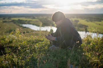 A teenage boy is sitting on the grass on a mountain against the backdrop of a beautiful sunset sky and using a mobile phone.